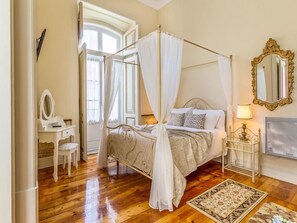 Una cama con dosel, una mesita de noche con lámpara, un espejo y una alfombra estampada sobre suelo de madera.