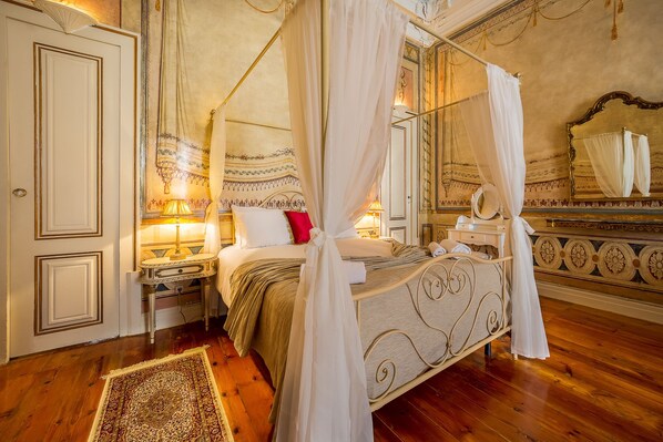 Una cama con dosel y cuatro columnas, con una mesita de noche y una lámpara, un espejo y una almohada roja.