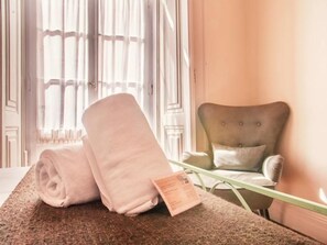 Una habitación con una cama, una toalla enrollada, una silla y un ventanal con cortinas.
