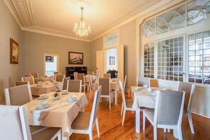 Un comedor con varias mesas dispuestas para el servicio, una araña y grandes ventanas con vidrieras decorativas.