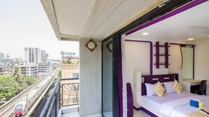 Quarto de hotel com varanda, cama com roupa de cama branca e almofadas amarelas, escrivaninha e vista para a cidade.
