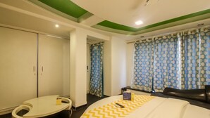 Quarto de hotel com cama, área de estar em curva, guarda-roupa e janela com cortinas estampadas.