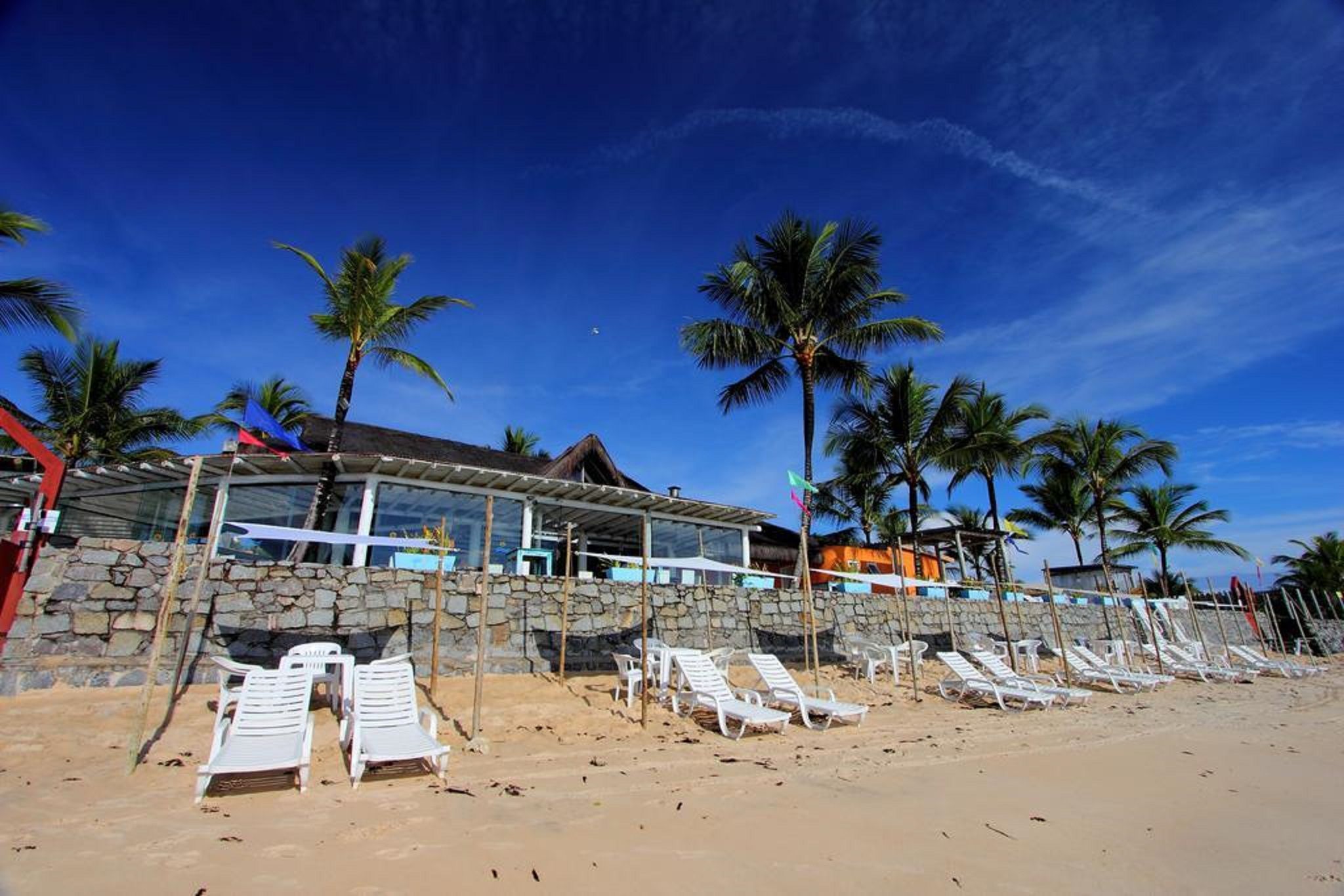 Um hotel à beira-mar com fachada de pedra, espreguiçadeiras brancas e palmeiras.