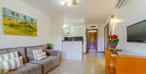 A living room with a sofa, a painting, a TV, and a kitchen in the background. - Mar Paraiso Hotel (Porto Seguro)