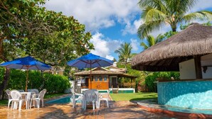 A pool area with white plastic chairs and tables, a thatched-roof structure, and a blue umbrella. - Mar Paraiso Hotel (Porto Seguro)