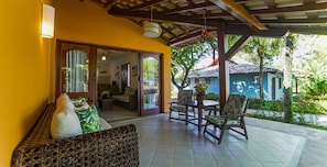 A covered outdoor seating area with a sofa and chairs, a dining table, and a view of a house and greenery. - Mar Paraiso Hotel (Porto Seguro)