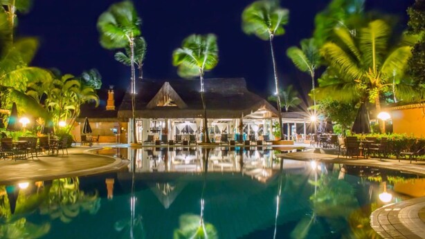 A nighttime view of a resort with a swimming pool reflecting palm trees and a building with a thatched roof. - Mar Paraiso Hotel (Porto Seguro)