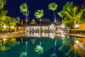 Vista nocturna de un complejo turístico con una piscina que refleja palmeras y un edificio con techo de paja.
