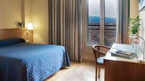 A hotel room with a bed, a desk with a chair, a lamp, a window with curtains, and a view of the cityscape. - Hotel J. Balmes Vic (Vic)
