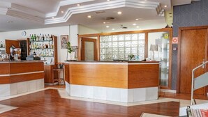 A hotel reception area with a wooden reception desk, a glass display case with bottles, and a clock on the wall. - Hotel J. Balmes Vic (Vic)