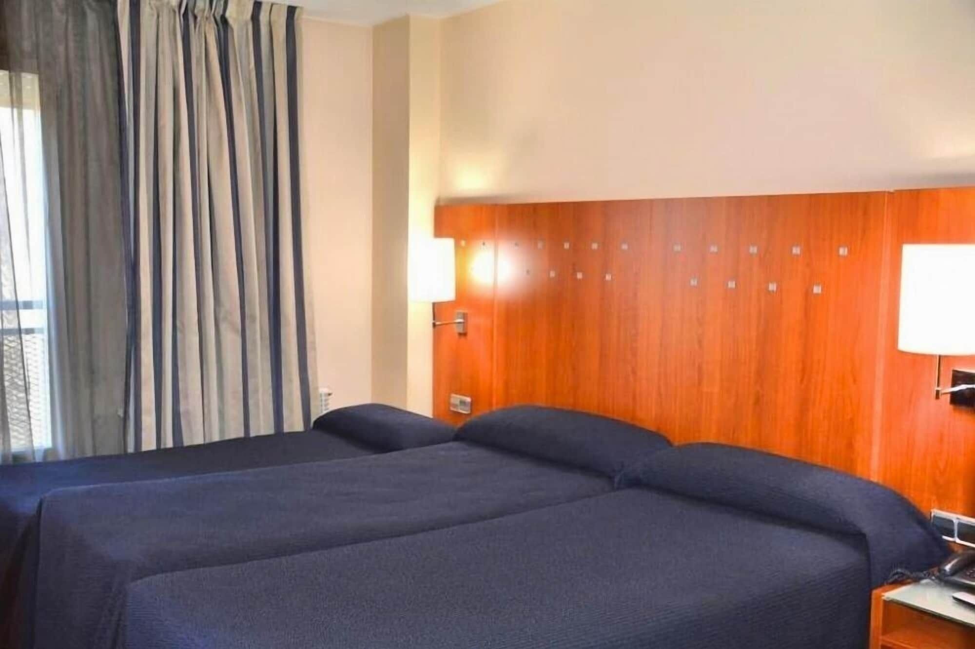Habitació d'hotel amb dos llits, un panell de fusta al capçal, una tauleta de nit amb una làmpada i una finestra amb cortines.