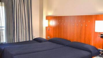 Habitació d'hotel amb dos llits, un panell de fusta al capçal, una tauleta de nit amb una làmpada i una finestra amb cortines.