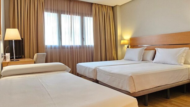 Een hotelkamer met twee bedden, een bureau met een lamp en een raam met gordijnen.