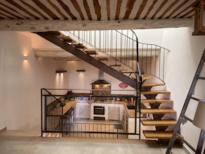 A modern kitchen with a central island, a built-in oven, and a stainless steel range hood. A spiral staircase with wooden steps and black metal railings leads to an upper level.