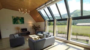 A spacious living room with a high ceiling, large windows offering a view of a lake and distant hills, and a modern chandelier. - House with breathtaking view of the mountains (Adast)