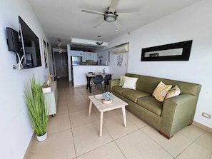 Uma sala de estar moderna com um sofá verde, uma mesa de centro de madeira e um ventilador de teto. Há uma área de jantar com cadeiras e uma mesa ao fundo.