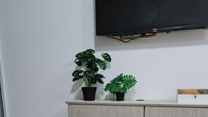 Una habitación con una mesa de madera, dos plantas en macetas, un jarrón azul con flores blancas y un televisor de pantalla plana montado en la pared.
