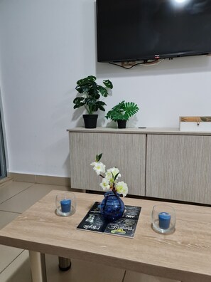 Une pièce avec une table en bois, deux plantes en pot, un vase bleu contenant des fleurs blanches et un téléviseur à écran plat fixé au mur.