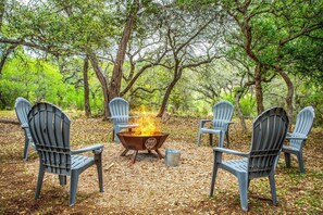 A campfire with four Adirondack chairs arranged around it in a forest setting. - The River Rock Retreat (Bandera)