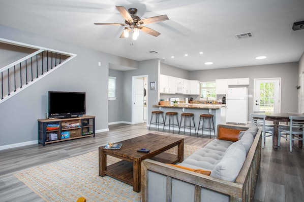 A spacious living room with a large sofa, a wooden coffee table, and a TV. There's a kitchen area with white cabinets and a bar. - The River Rock Retreat (Bandera)