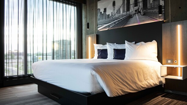 Quarto de hotel moderno com uma cama grande, vista para a cidade e uma moldura com uma imagem de uma ponte.