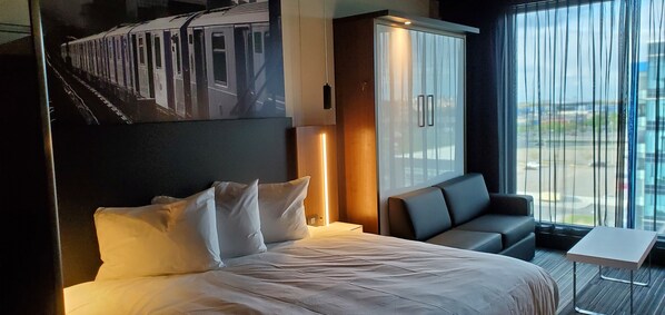 Habitación de hotel con cama, sofá, una mesita y un ventanal con vistas al exterior.