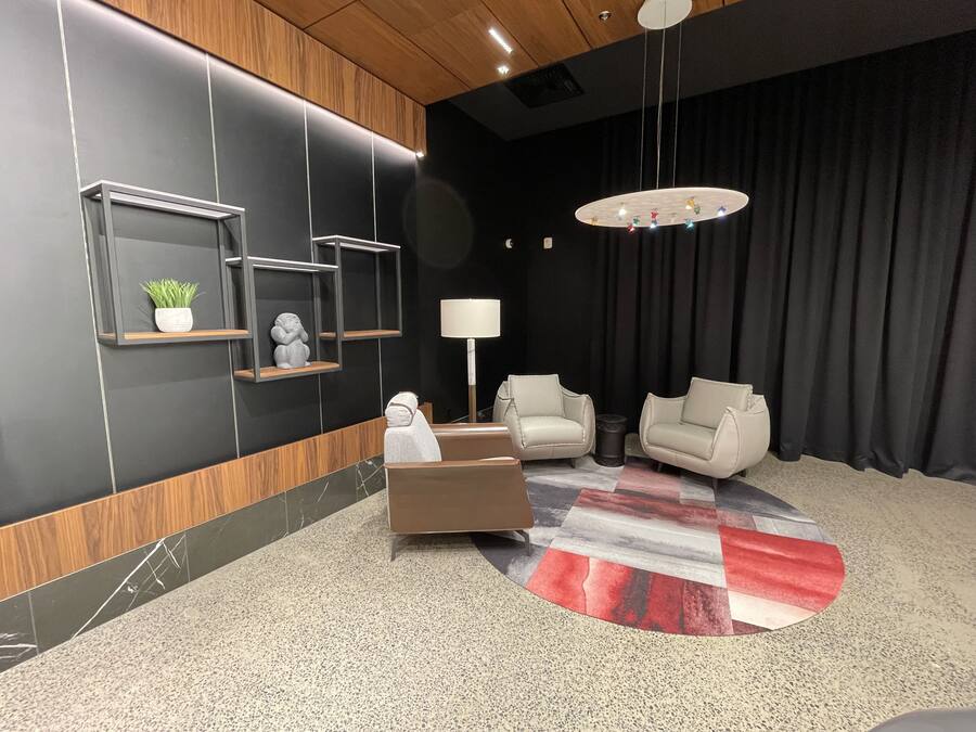 A modern lobby with a black and wood panel wall, a marble floor, a central brown sofa, two white armchairs, a round rug, and a hanging light fixture.