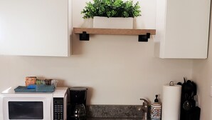 A kitchen with white cabinets, a stainless steel sink, a microwave, a coffee maker, and a shelf with a plant.
