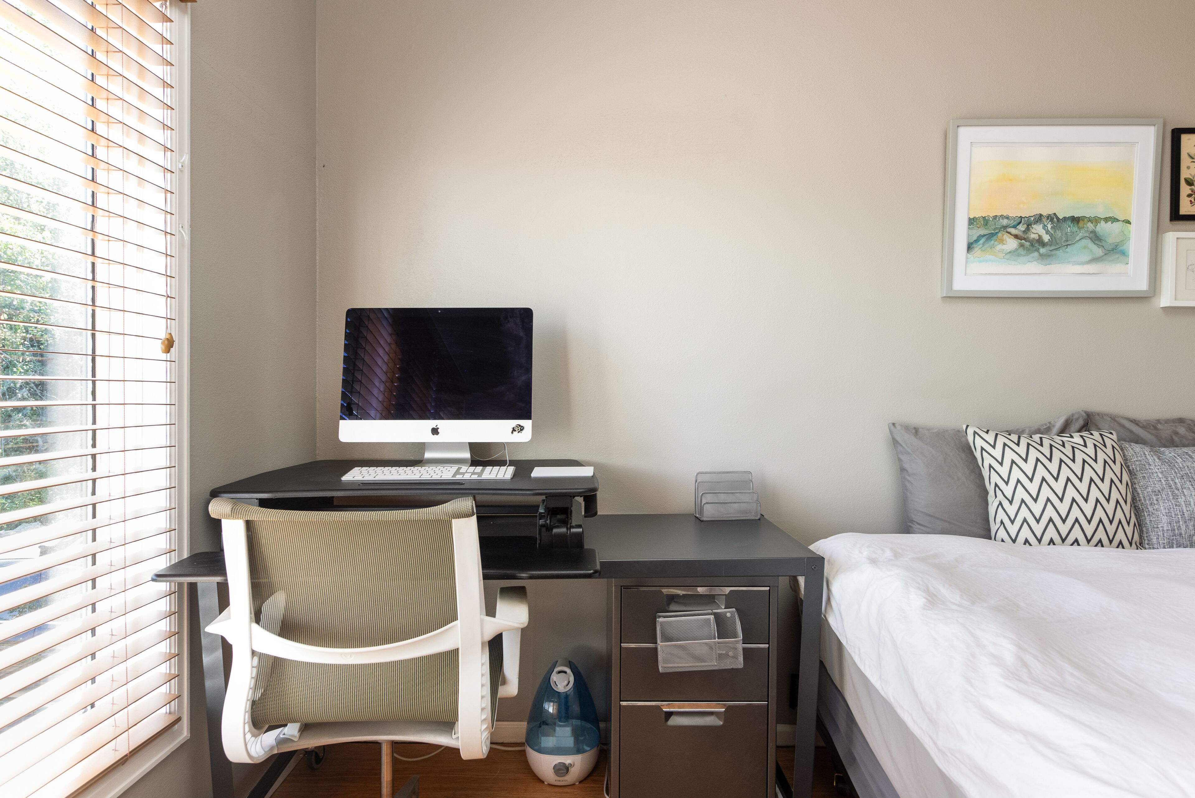 A bedroom with a bed, a desk with a computer, a chair, a window with blinds, and a wall-mounted artwork.