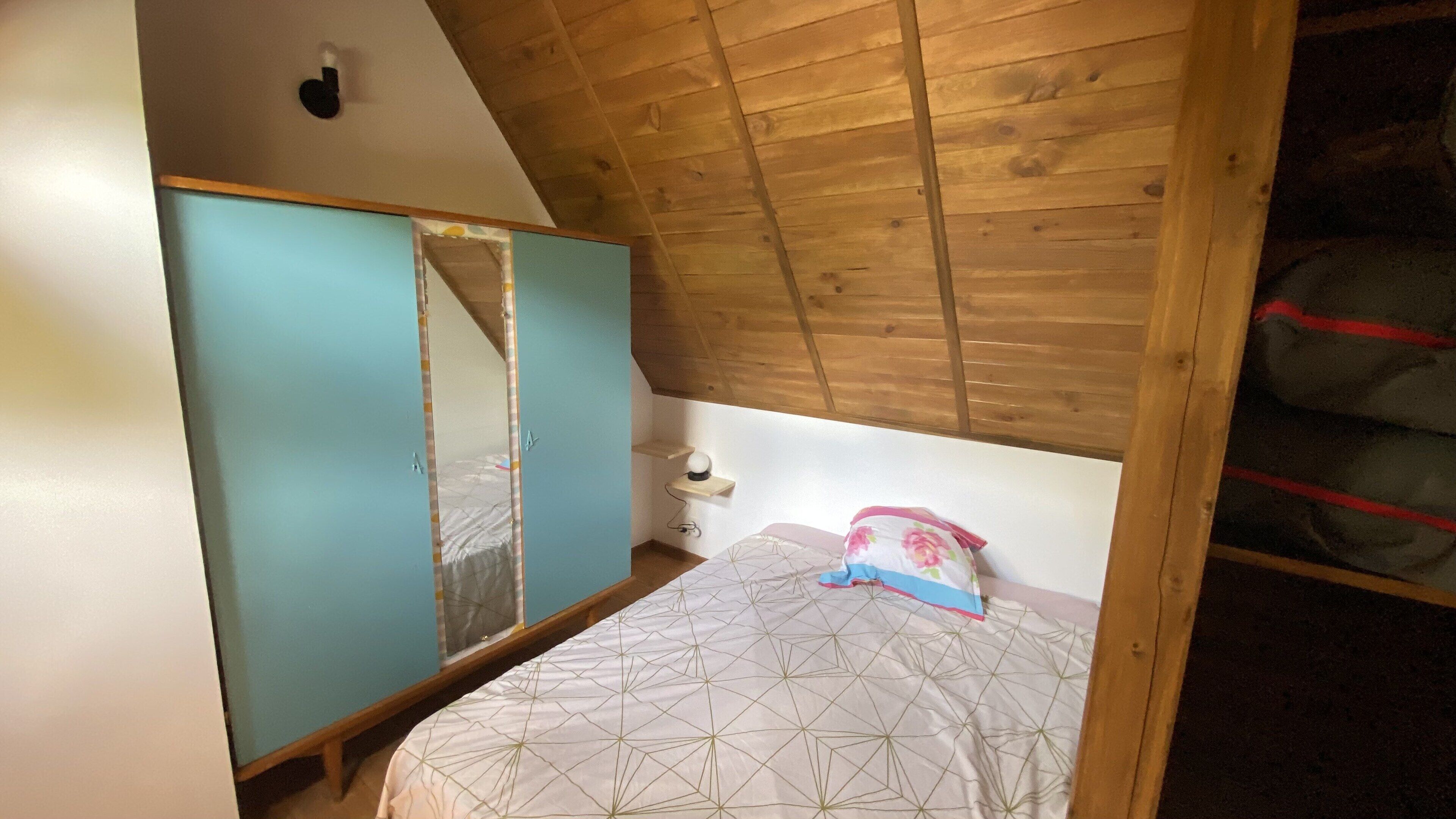 Un dormitorio con techo inclinado, una cama con colcha estampada, una mesita de noche con lámpara y un armario.