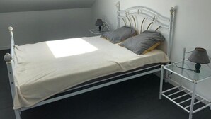 Una cama individual con estructura metálica blanca, dos almohadas, una mesita de noche con lámpara y una pequeña mesa con lámpara.