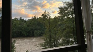 Une vue sur une rivière bordée d’une forêt dense, aperçue à travers une fenêtre ornée d’un rideau.