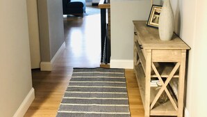 Un pasillo con piso de madera, un tapete estampado, una consola de madera con un jarrón y un espejo en la pared.
