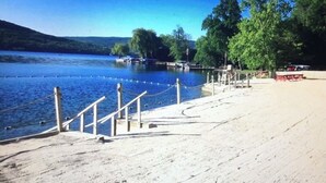 Una playa arenosa con una escalera de madera que lleva a un muelle, un lago azul cristalino y árboles al fondo.