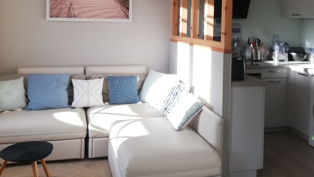Una sala de estar moderna con un sofá blanco, una gran fotografía de playa en la pared y una zona de cocina con armarios blancos.