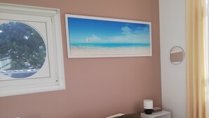 Un dormitorio con una cama, dos mesitas de noche, una lámpara de pared, un cuadro enmarcado de una playa y una ventana con vista a árboles.