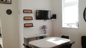 Un comedor moderno con una mesa blanca, sillas negras, un televisor montado y una ventana con persianas.