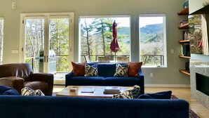 Una sala de estar con un sofá azul, un sillón de cuero marrón y una mesa de centro. Hay dos ventanas amplias con vistas a árboles y colinas.