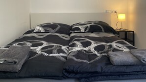 Una cama con una colcha estampada en blanco y negro, una almohada y una toalla doblada. Encima de la cama, hay un cuadro con la palabra “ADVENTURE” en letras rosas.