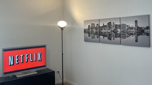 Una sala de estar con un televisor de pantalla plana mostrando 'NETFLIX', una lámpara de pie y una obra de arte con un paisaje urbano en la pared.