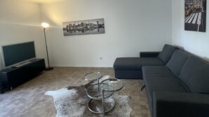Una sala de estar moderna con un sofá modular, un televisor de pantalla plana, una mesa de centro, una lámpara de pie y una pintura con vista a un paisaje urbano.