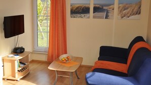 Ein Wohnzimmer mit Fernseher, Radio, einem kleinen Tisch mit Obstschale, einer Couch und drei Bildern mit Strandmotiv an der Wand.