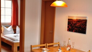 Ein Essbereich mit Holztisch und Stühlen, einer Hängelampe, einem Bild an der Wand und einem Bett im angrenzenden Zimmer.