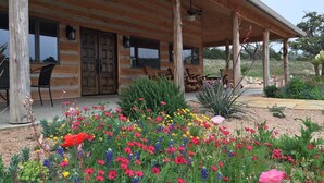 A wooden cabin with a covered porch, outdoor seating, and a garden with colorful flowers. - Hill Country Ranch Retreat (Johnson City)