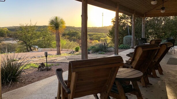 A covered patio with wooden chairs and tables, overlooking a landscaped garden with various plants and a distant view of hills. - Hill Country Ranch Retreat (Johnson City)