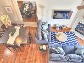 A modern living room with a dining area, grey sofa, blue patterned rug, and a flat-screen TV mounted on the wall.