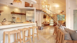 Une cuisine moderne dotée d’un îlot central, de tabourets en bois, d’un escalier et d’un espace de vie comprenant un canapé et une lampe.