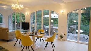 A spacious living room with a modern chandelier, a sofa, and a dining area with yellow chairs and a table. Large windows offer views of the outdoors. - Luxury apartment in the olive grove in the hinterland of Alassio/Albenga, carport, Wifi (Casa nova Lerrone)