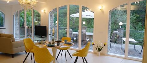 Amplia sala de estar con una lámpara moderna, un sofá y una zona de comedor con sillas amarillas y una mesa. Grandes ventanales ofrecen vistas al exterior.