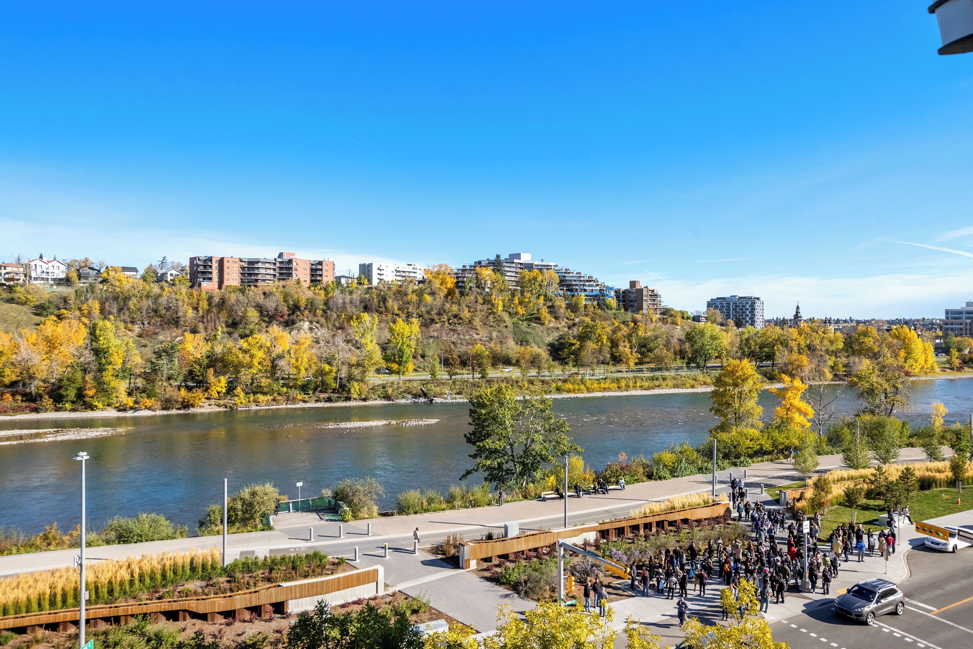 A riverside walkway with a crowd, a bridge, and buildings in the background.